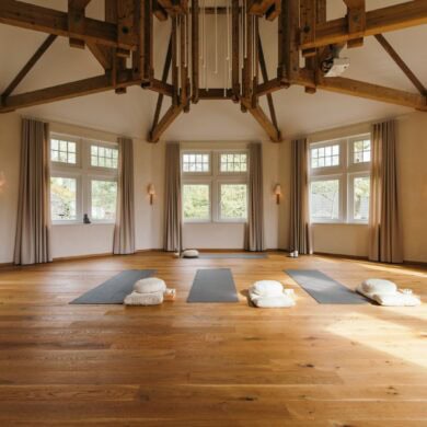 In einem achteckigen Raum mit weißen Wänden und großen Fenstern liegen drei Yogamatten mit Zubehör auf dem hölzernen Dieleboden. An der weißen Decke ist ein parallel zur Raumform laufendes Gebälk angebracht