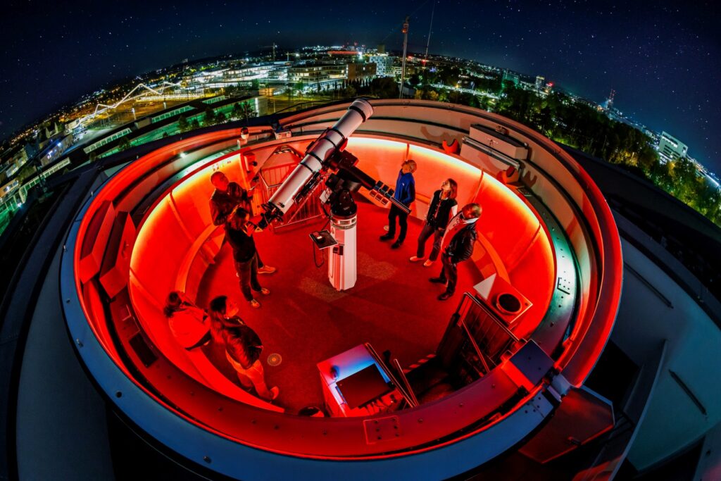 Die Experimenta Sternenwarte während der Skynights, von oben aufgenommen. In einem geöffneten, rot beleuchteten Observatorium stehen mehrere Menschen um ein zentrales Teleskop herum und schauen in den Himmel.