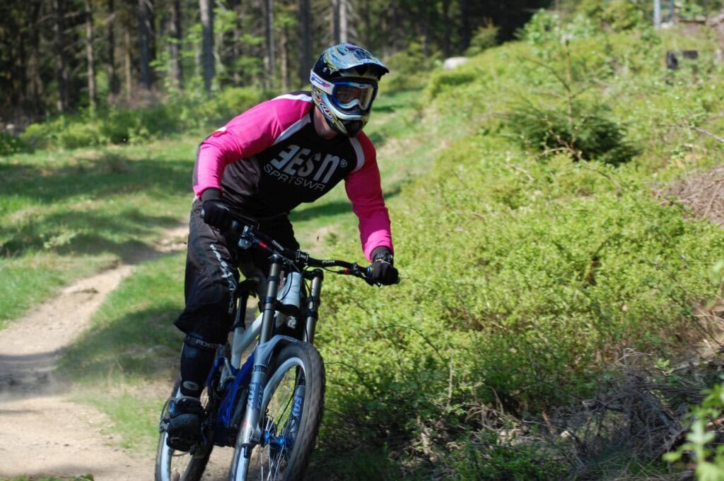 Ein Mann in Sportkleidung, Fahrradhelm und großer Sportbrille fährt stehend auf einem Mountainbike einen Trail entlang. Im Hintergrund sieht man den Weg, der geschlängelt aus einem Waldstück herausläuft