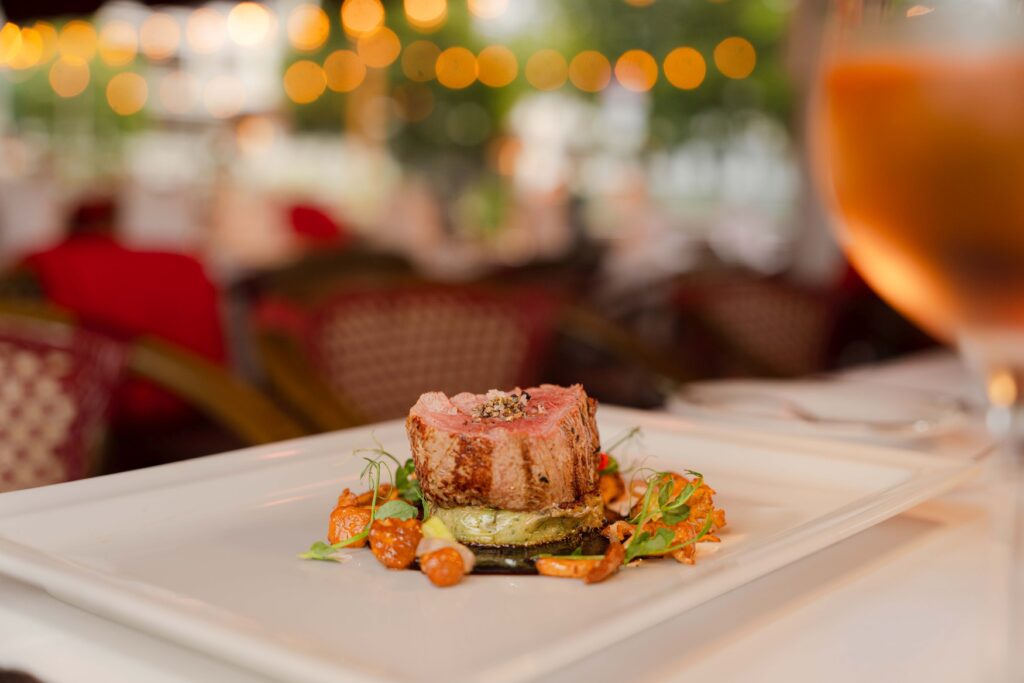 A pink piece of veal fillet is served on a square plate on a bed of fried chanterelle mushrooms, garnished with a few herbs. A glass of rosé wine is blurred in the foreground.