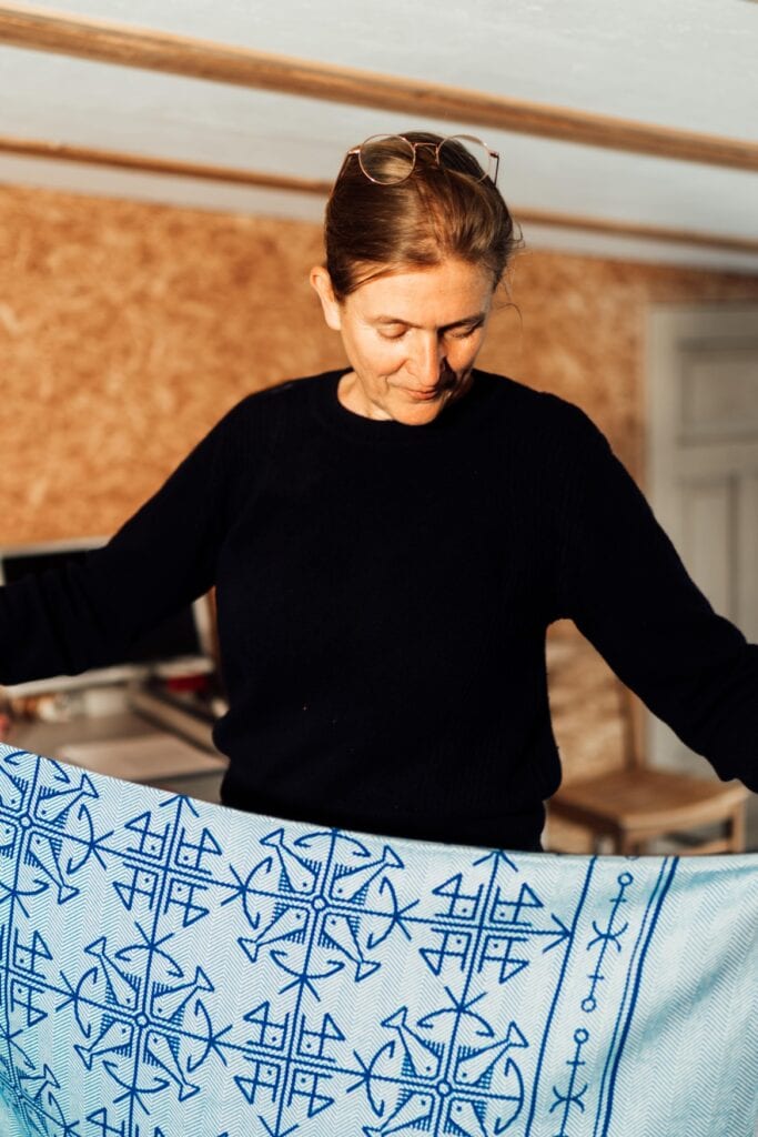 Eine Frau mittleren Alters mit rötlichem, zurückgebundenen Haar, in das eine Brille geschoben ist, und einem schwarzen Pulli hält einen grau-blauen Teppich mit geometrischen Mustern in Händen und blickt darauf runter.