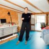 Eine Frau mittleren Alters mit rötlichem Haar und schwarzer Freizeitkleidung steht, die Arme verschränkt, in einem Atelier. Sie blickt direkt in die Kamera. Links von ihr ein Arbeitstisch mit breiten, flachen Schubladen, rechts ein Tisch, über dessen Rand ein blauer Teppich hängt. Die Wände sind mit Spanholz verkleidet, der Boden mit blauen Linoleumplatten ausgelegt.