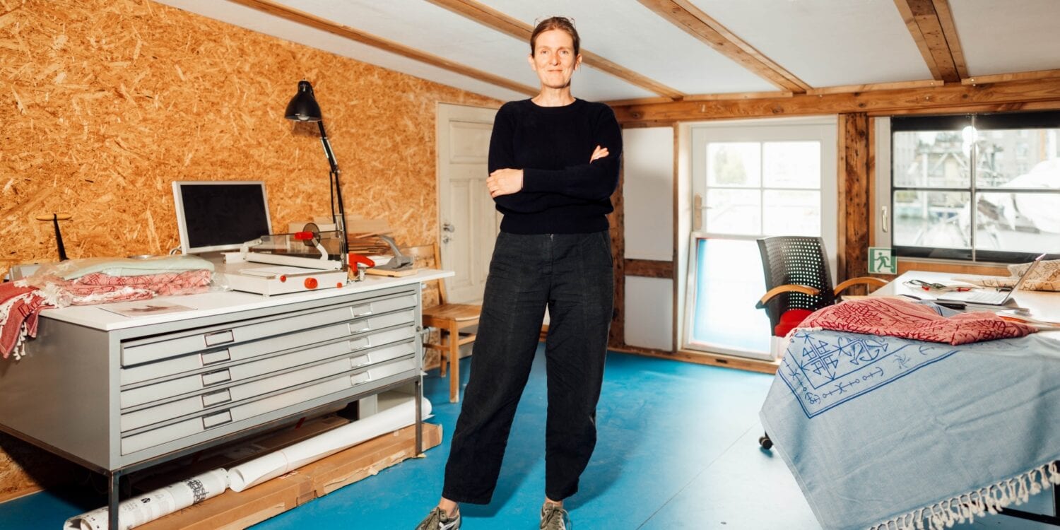 Eine Frau mittleren Alters mit rötlichem Haar und schwarzer Freizeitkleidung steht, die Arme verschränkt, in einem Atelier. Sie blickt direkt in die Kamera. Links von ihr ein Arbeitstisch mit breiten, flachen Schubladen, rechts ein Tisch, über dessen Rand ein blauer Teppich hängt. Die Wände sind mit Spanholz verkleidet, der Boden mit blauen Linoleumplatten ausgelegt.