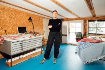 Eine Frau mittleren Alters mit rötlichem Haar und schwarzer Freizeitkleidung steht, die Arme verschränkt, in einem Atelier. Sie blickt direkt in die Kamera. Links von ihr ein Arbeitstisch mit breiten, flachen Schubladen, rechts ein Tisch, über dessen Rand ein blauer Teppich hängt. Die Wände sind mit Spanholz verkleidet, der Boden mit blauen Linoleumplatten ausgelegt.