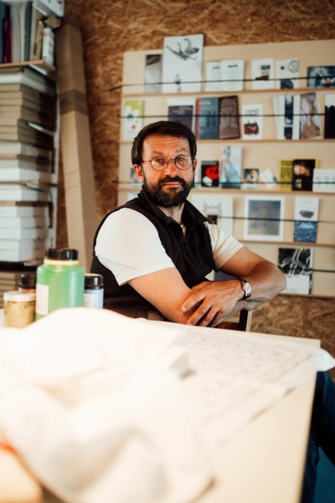 Ein Mann mittleren Alters, mit schwarzem Haar und Bart und einer halbrunden Metallbrille, sitzt in weißem T-Shirt und schwarzer Weste mit dem Körper leicht abgewandt hinter einem Tisch, auf dem unscharf erkennbar einige Schnittmuster liegen. Er blickt direkt in die Kamera. Hinter ihm ein flaches Regal mit mehreren Prospekten und Flyern