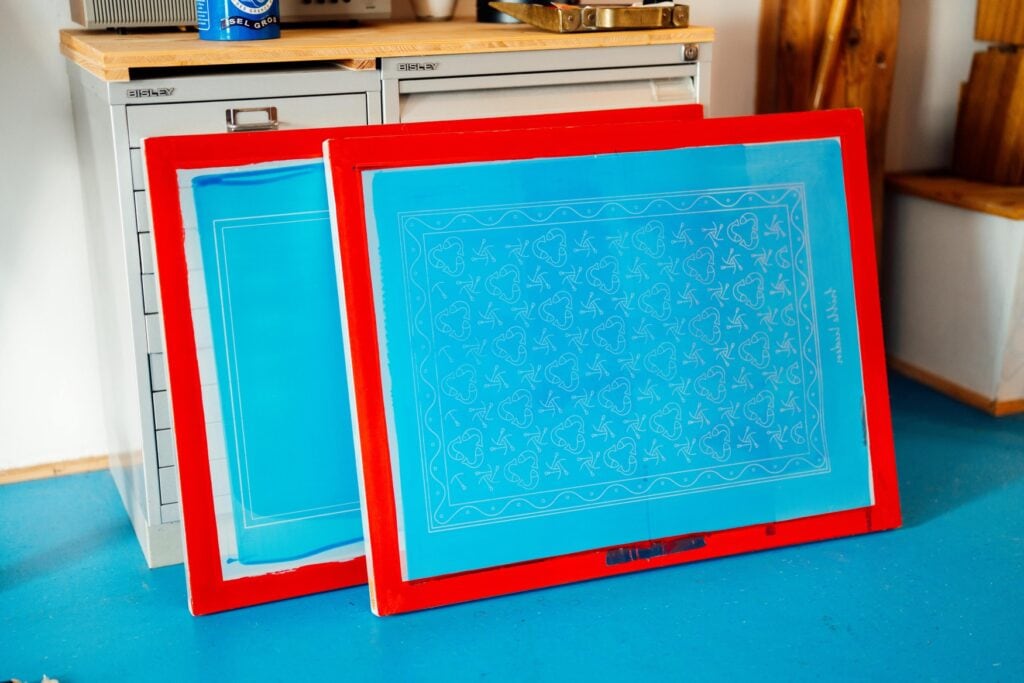 Vor einem hüfthohen, blauen Werkschrank lehnen zwei Siebdruckplatten. Darauf sind weiße Muster erkennbar, die Platten selbst wurden mit blauer Farbe bestrichen, der Rand wiederum jeweils in kräftigem Rot.