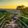 A rocky hiking trail, bordered by metal railings, leads to a bench on a hilltop, centered in the picture. To the left and right of the trail are a few small trees. Behind them, the view opens up to a low mountain range with a small town, all bathed in the golden light of sunset.