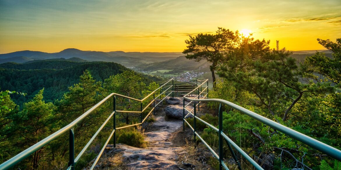 A rocky hiking trail, bordered by metal railings, leads to a bench on a hilltop, centered in the picture. To the left and right of the trail are a few small trees. Behind them, the view opens up to a low mountain range with a small town, all bathed in the golden light of sunset.