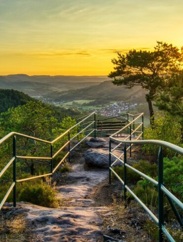 A rocky hiking trail, bordered by metal railings, leads to a bench on a hilltop, centered in the picture. To the left and right of the trail are a few small trees. Behind them, the view opens up to a low mountain range with a small town, all bathed in the golden light of sunset.
