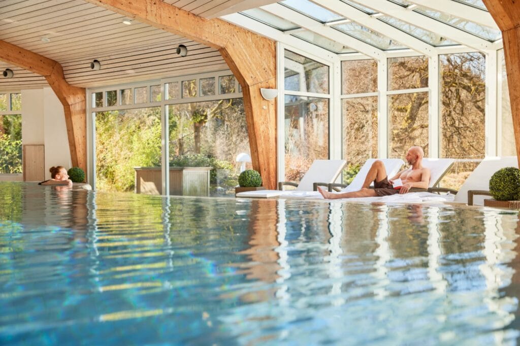 Die Hälfte des Bildes wird von einem Pool eingenommen, in dem links eine Frau im Wasser auf dem Beckenrand lehnt. Das Schwimmbad hat viele bodentiefe Fenster und helle Holzbalken, wodurch alles sehr lichtdurchflutet wirkt. Auf der rechten Seite am Rand liegt ein Mann auf einer Liege.