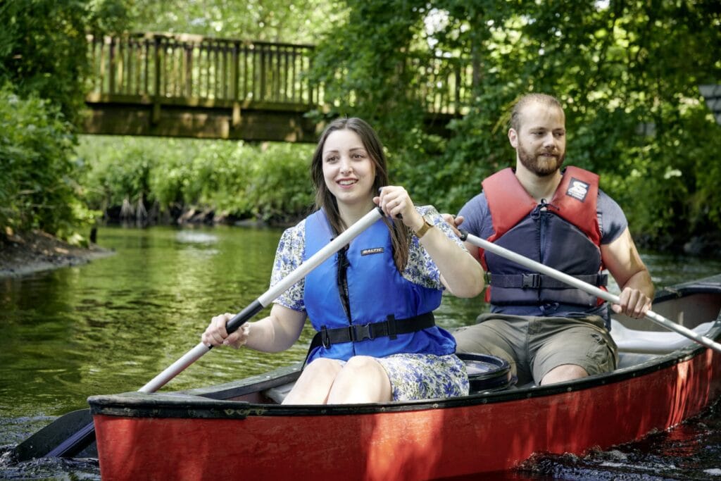Eine junge Frau und ein junger Mann in sommerlicher Kleidung und je mit Schwimmweste paddeln in einem roten Kanu auf einem Fluss. Das Ufer ist bewachsen, im Hintergrund spannt eine kleine Brücke über den Fluss.