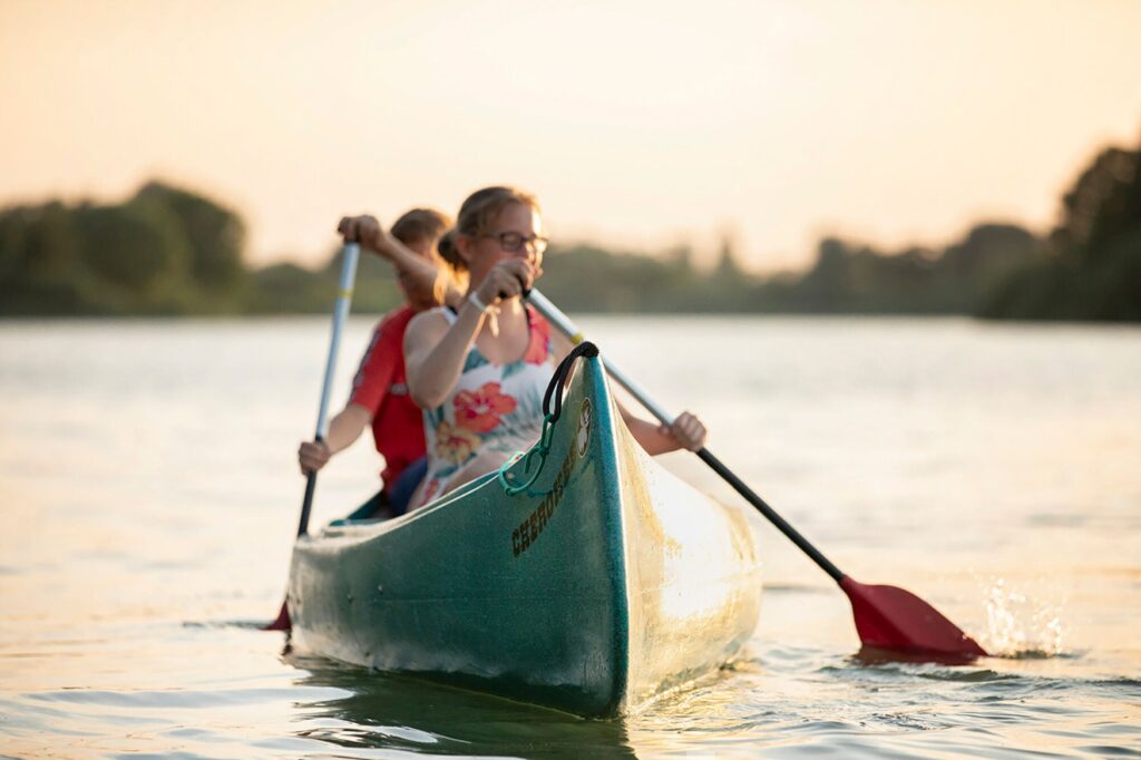 Zwei junge Menschen paddeln auf einem grünen Kanu auf einem großen See oder breiten Fluss im Licht des Sonnenuntergangs. Im Hintergrund ist unscharf das grüne Ufer zu erkennen.