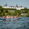 Ein großes Kanu mit zehn Personen in Schwimmwesten fährt auf einem Gewässer von links nach rechts. Im Hintergrund ist das grüne Ufer erkennen, dahinter eine Anhöhe mit einem prächtigen, weißen Schloss darauf. Auf dem Kanu steht der Schriftzug "Kanu-Stadtführung"