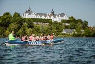 Ein großes Kanu mit zehn Personen in Schwimmwesten fährt auf einem Gewässer von links nach rechts. Im Hintergrund ist das grüne Ufer erkennen, dahinter eine Anhöhe mit einem prächtigen, weißen Schloss darauf. Auf dem Kanu steht der Schriftzug "Kanu-Stadtführung"