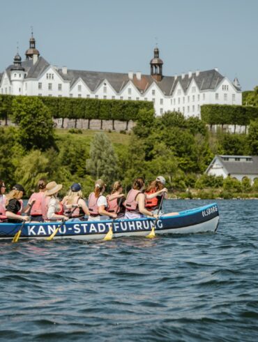 Ein großes Kanu mit zehn Personen in Schwimmwesten fährt auf einem Gewässer von links nach rechts. Im Hintergrund ist das grüne Ufer erkennen, dahinter eine Anhöhe mit einem prächtigen, weißen Schloss darauf. Auf dem Kanu steht der Schriftzug "Kanu-Stadtführung"