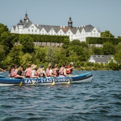 Ein großes Kanu mit zehn Personen in Schwimmwesten fährt auf einem Gewässer von links nach rechts. Im Hintergrund ist das grüne Ufer erkennen, dahinter eine Anhöhe mit einem prächtigen, weißen Schloss darauf. Auf dem Kanu steht der Schriftzug "Kanu-Stadtführung"