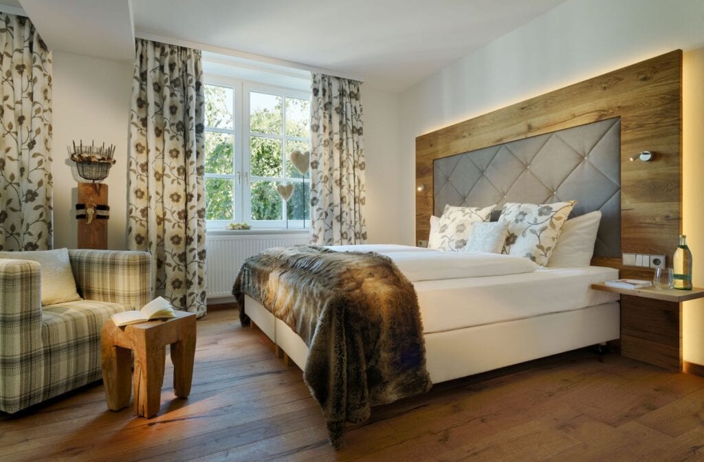 Ein Hotelzimmer, rechts das Bett mit Lederkopfteil, das in eine hölzerne Wandverkleidung eingelassen ist, mittig ein Doppelfenster mit Heizung darunter, links ein eckiger, grün-weiß karierter Sessel vor einem kleinen Holzbeistelltisch. Die Wände sind weiß, der Boden besteht aus Holzdielen