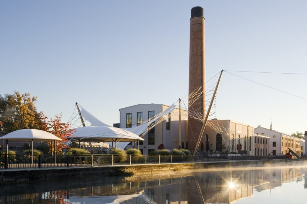 Ein altes, aber renoviertes Fabrikgebäude mit hohem Schornstein steht an einem Kanal, in dessen Wasser sich die Abendsonne spiegelt. Links vom Gebäude stehen zwei runde Pavillons und einige Bäume, eine Uferpromenade ist durch einen Zaun zum Kanal abgegrenzt.