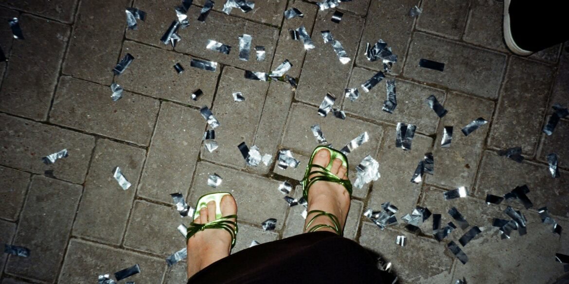 Ein Paar Füße in grün-glänzenden High-Heels stehen auf Pflastersteinen, rundherum silbernes Konfetti. Das Foto sieht aus, wie von der Fußinhaberin selbst fotografiert.