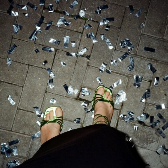 Ein Paar Füße in grün-glänzenden High-Heels stehen auf Pflastersteinen, rundherum silbernes Konfetti. Das Foto sieht aus, wie von der Fußinhaberin selbst fotografiert.