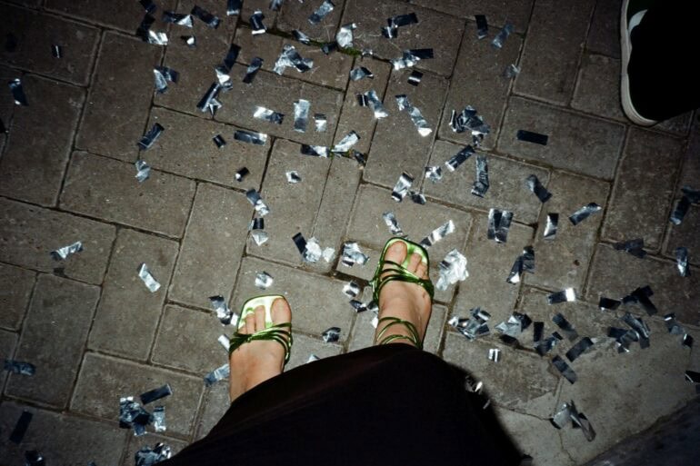 Ein Paar Füße in grün-glänzenden High-Heels stehen auf Pflastersteinen, rundherum silbernes Konfetti. Das Foto sieht aus, wie von der Fußinhaberin selbst fotografiert.