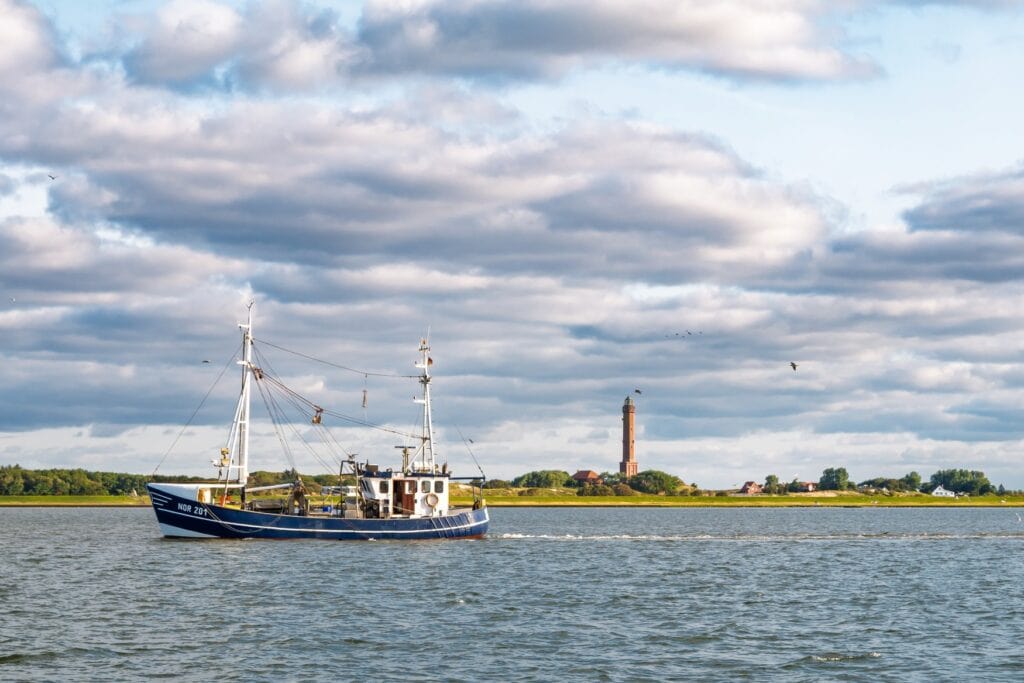 Ein kleiner blauer Fischkutter fährt auf dem Meer von rechts nach links durchs Bild. Im Hintergrund eine flache, grüne Insel mit etwas Waldbewuchs, mittig ragt ein Backsteinleuchtturm in den Himmel