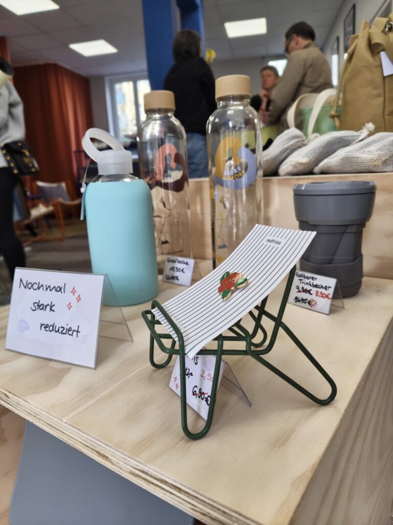 Display in a fair fashion store: A rose-shaped pin is draped over a miniature folding chair, with various reusable cups and bottles behind it. All products are labeled with prices on small signs. Several customers can be seen blurred in the background.
