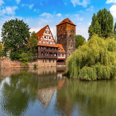 A wide river, flowing from left to right. On the left, the river is spanned by a three-arched sandstone bridge; opposite the viewer is a wide quay wall with a multi-story half-timbered house in the center and an angular stone tower next to it. On the right, next to a large weeping willow, is a wooden bridge with a roof.
