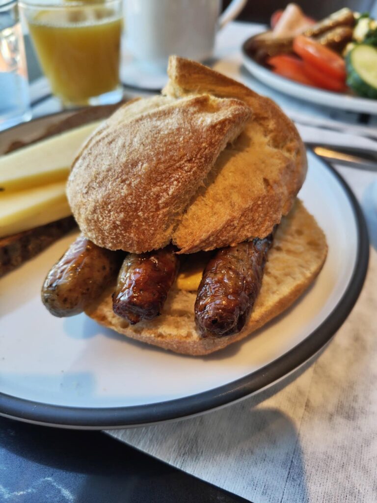 Close-up of a roll with three Nuremberg sausages on a plate; behind it, another plate with sausages, tomatoes, and cucumbers