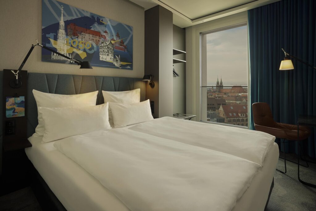 A double bed with two sets of blankets and pillows in a hotel room; above the bed hangs a modern collage print featuring various sketches of buildings in Nuremberg and Dürer's hare. To the right behind the bed, a floor-to-ceiling window opens up to a view over the rooftops of Nuremberg.