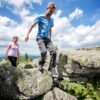 Ein Mann und eine Frau in sommerlicher Outdoorkleidung steigen über einige Felsbrocken auf einer Bergkuppe. Auf einem der Felsen ist ein mit weißer Kreide nachgezogener Ochsenkopf eingeritzt. Im Hintergrund sieht man das Tal mit vielen Wäldern. Über allem blauer Himmel mit ein paar Wolken.