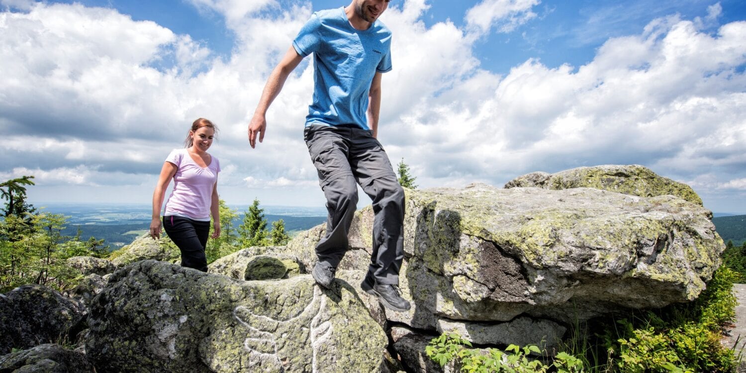 Ein Mann und eine Frau in sommerlicher Outdoorkleidung steigen über einige Felsbrocken auf einer Bergkuppe. Auf einem der Felsen ist ein mit weißer Kreide nachgezogener Ochsenkopf eingeritzt. Im Hintergrund sieht man das Tal mit vielen Wäldern. Über allem blauer Himmel mit ein paar Wolken.