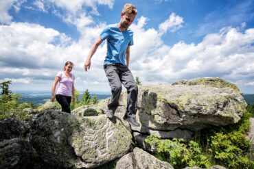 Ein Mann und eine Frau in sommerlicher Outdoorkleidung steigen über einige Felsbrocken auf einer Bergkuppe. Auf einem der Felsen ist ein mit weißer Kreide nachgezogener Ochsenkopf eingeritzt. Im Hintergrund sieht man das Tal mit vielen Wäldern. Über allem blauer Himmel mit ein paar Wolken.