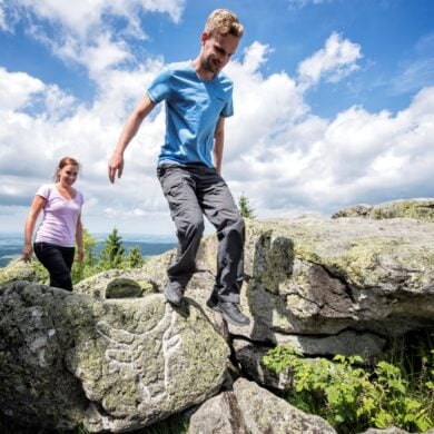 Ein Mann und eine Frau in sommerlicher Outdoorkleidung steigen über einige Felsbrocken auf einer Bergkuppe. Auf einem der Felsen ist ein mit weißer Kreide nachgezogener Ochsenkopf eingeritzt. Im Hintergrund sieht man das Tal mit vielen Wäldern. Über allem blauer Himmel mit ein paar Wolken.