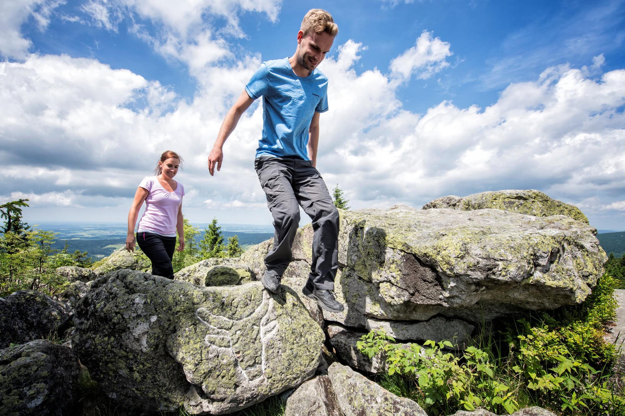 Ein Mann und eine Frau in sommerlicher Outdoorkleidung steigen über einige Felsbrocken auf einer Bergkuppe. Auf einem der Felsen ist ein mit weißer Kreide nachgezogener Ochsenkopf eingeritzt. Im Hintergrund sieht man das Tal mit vielen Wäldern. Über allem blauer Himmel mit ein paar Wolken.