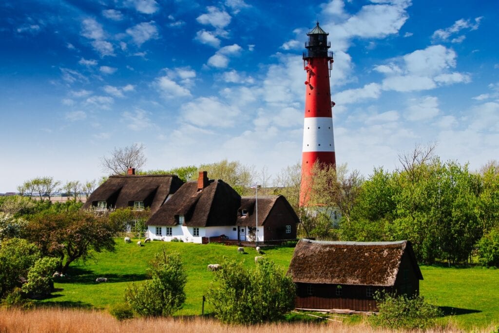 Inmitten einiger typisch nordfriesischer Häuser mit weißem Putz und braunem Reetdach ragt ein rot-weiß gestreifter in den blauen Himmel. Um die Häuser herum grasen einige Schafe auf einer Wiese.