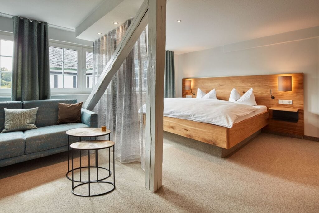 Ein Hotelzimmer, mittig durch grau gestrichenes, frei stehendes Gebälk getrennt. Hinter dem Gebälk das Bett mit Holzrahmen und -kopfstück, vor dem Gebälk auf der linken Seite eine grau-grüne Zweiercouch, der Boden mit Teppich ausgekleidet. Durch ein wandbreites Fenster fällt Tageslicht hinein.