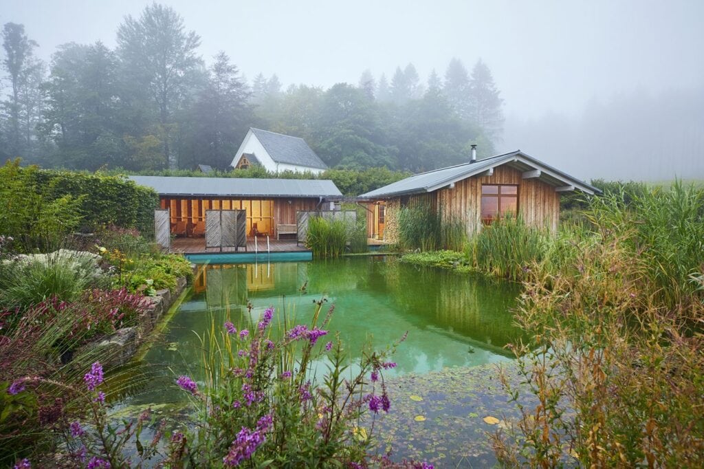 Um einen eckigen Outdoorpool, dessen Rand dicht bewachsen ist, läuft hinten und rechts eine hölzerne, eingeschossige Hütte, vor der eine kleine Veranda angebracht ist. Hütte und Veranda sind in ein gemütliches, gelbes Licht getaucht. Dahinter ragt aus dichtem Nebel etwas Wald hervor.