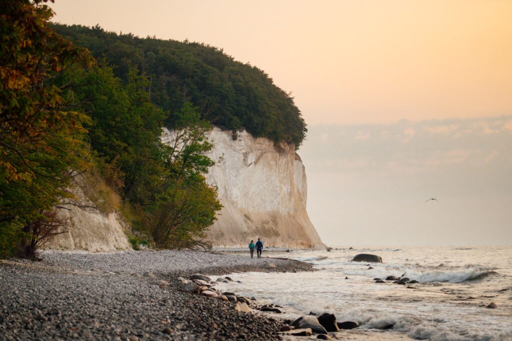Two people are walking in front of the chalk cliffs on Rügen, which rise from the center of the image to the left. On the right, the Baltic Sea borders the beach, and everything is bathed in the light of the setting sun.