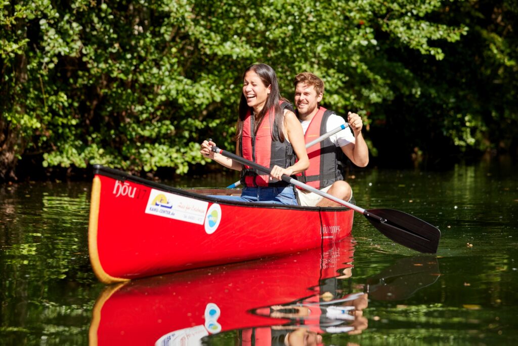 Ein junger Mann und eine junge Frau auf einem roten Kanu im Wasser bei strahlender Sonne. Beide lachen. Im Hintergrund dichter Uferbewuchs