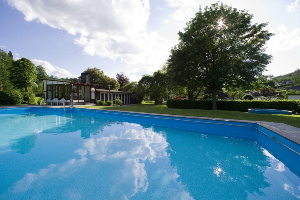Ein großer Swimmingpool im Freien. Der Rand dahinter grenzt an einen grünen Rasen, rechts dahinter eine kleine Hecke und ein großer Laubbaum. Links im Bild hinter dem Pool noch ein eingeschossiges Gebäude mit bodentiefen Fenstern und Fachwerkdekor.