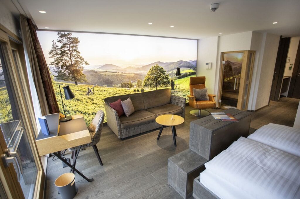 Ein Hotelzimmer, mit grauen Holzdielen, einem grauen Bett in der rechten Ecke, einem wandhohen Fenster, das den Blick auf eine grüne Hügellandschaft öffnet. Vor dem Fenster stehen ein graues Zweiersofa und rechts davon ein moderner, brauner Ledersessel. Links im Bild noch ein kleiner Holzschreibtisch.