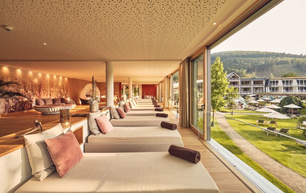 Eine Ruhebereich in einem Spa. Links ein hölzernes Plateu, rechts daran angrenzend und etwas abgesenkt ein langer Bereich mit mehreren Liegen, die auf eine breite Fensterwand auf der rechten Seite ausgerichtet sind. Außen ist eine grüne Wiese mit einigen Liegestühlen, weiter im Hintergrund ein dreistöckiges Hotelgebäude, hinter dem die grünen Hügel des Sauerlandes auftauchen.