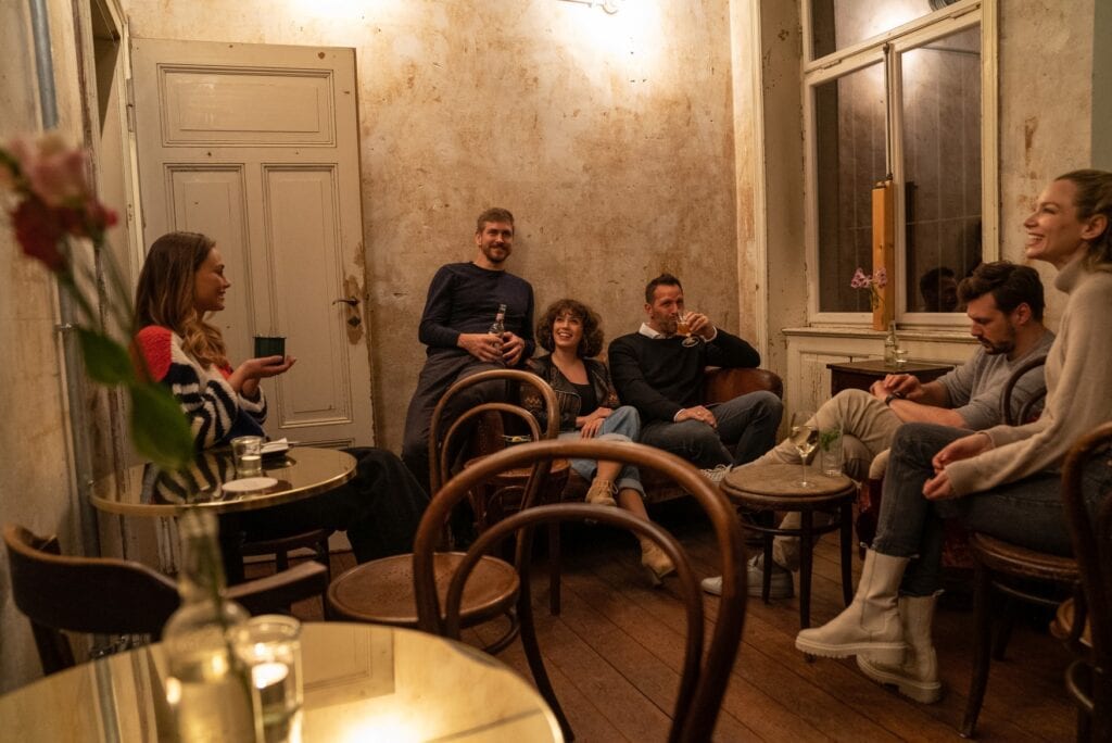In einem kleinen Altbau-Raum sitzen mehreere Menschen mit verschiedenen Getränken auf Holzstühlen und einem Sofa. Die Stimmung scheint locker, durch das Fenster wird erkennbar, dass es Nacht ist. Von der Decke leuchtet ein gemütlich-gelbliches Licht.