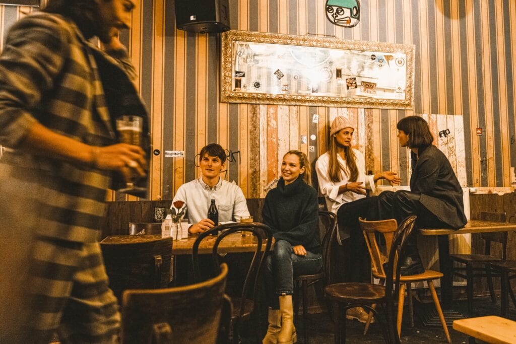 Gästeraum in einer Bar. An einem eckigen Tisch sitzen ein Mann und eine Frau und schauen lächelnd auf einen weiteren Mann in einem karierten Anzug, der vor ihnen vorbeigeht. Rechts von ihnen lehnt eine Frau an der Wand und unterhält sich mit einer weiteren Frau, die ihr zugewandt auf einem Tisch sitzt, die Füße auf einem Stuhl abgelegt.