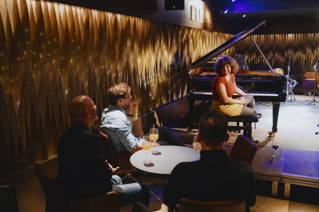 Szene in einem Jazzclub. Drei Männer sitzen mit dem Rücken zur Kamera an einem runden Café-Tisch, darauf drei Weingläser. Sie unterhalten sich mit einer Frau auf der Bühne vor ihnen, die an einem Flügel sitzt und sich lachend zur Dreiergruppe zuwendet. Das Licht in dem Raum ist gemütlich gedimmt, links an der Wand sind gelb-goldene Panele angebracht.