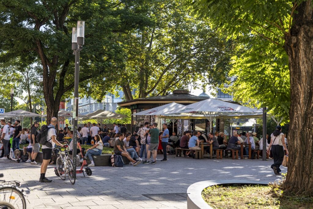 Ein belebter Platz in einer Stadt. Unter mehreren Sonnenschirmen sitzen Menschen an Bierzeltgarnituren oder auf kleinen Mäuerchen. Einige hohe Bäume spenden Schatten. Hinter einigen Sonnenschirmen ist ein Pavillon bzw. ein Milchhäuschen zu sehen.