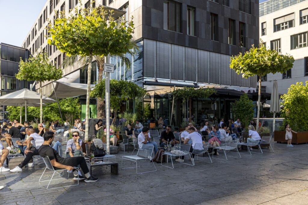 Auf einem Platz vor einem modernen, mit dunklen Metallplatten verkleideten Gebäude sitzen viele Menschen in sommerlicher Kleidung in einer Außengastronomie. Teilweise fällt der Schatten des Gebäudes auf die Szenerie.