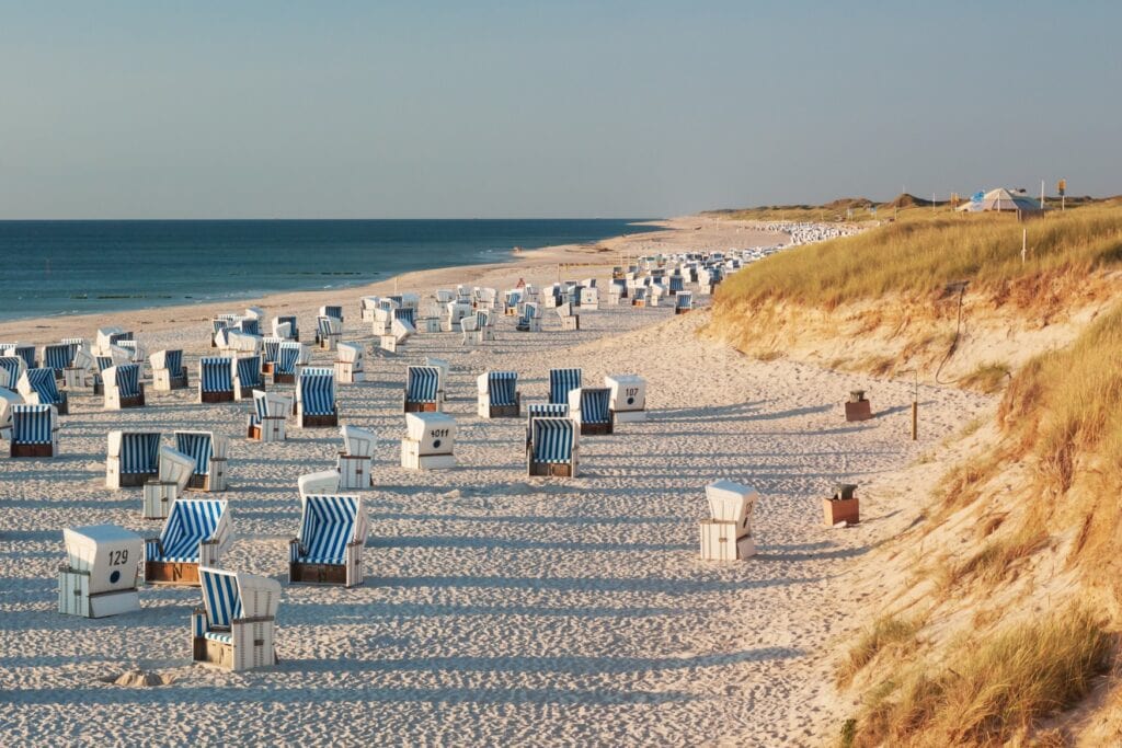 Auf einem langezogenen Strand stehen zahllose, weiß-blau gestreifte Strandkörbe nebeneinander im Licht der untergehenden Sonne. Links grenzt das Meer an den Strand, rechts eine bewachsene Düne.