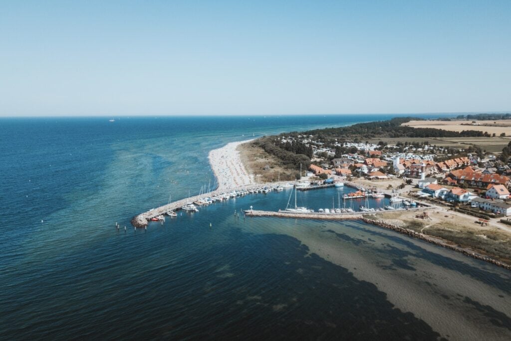 Luftaufnahme einer Insel; die Insel liegt auf der rechten Seite des Bildes, umgeben von Meer. Im Fokus ein kleiner Hafen mit mehreren Segelbooten und kleineren Yachten, an den sich ein kleiner Ort mit rotgedeckten Häusern anschließt. Hinter dem Ort grenzt ein kleiner Wald an.